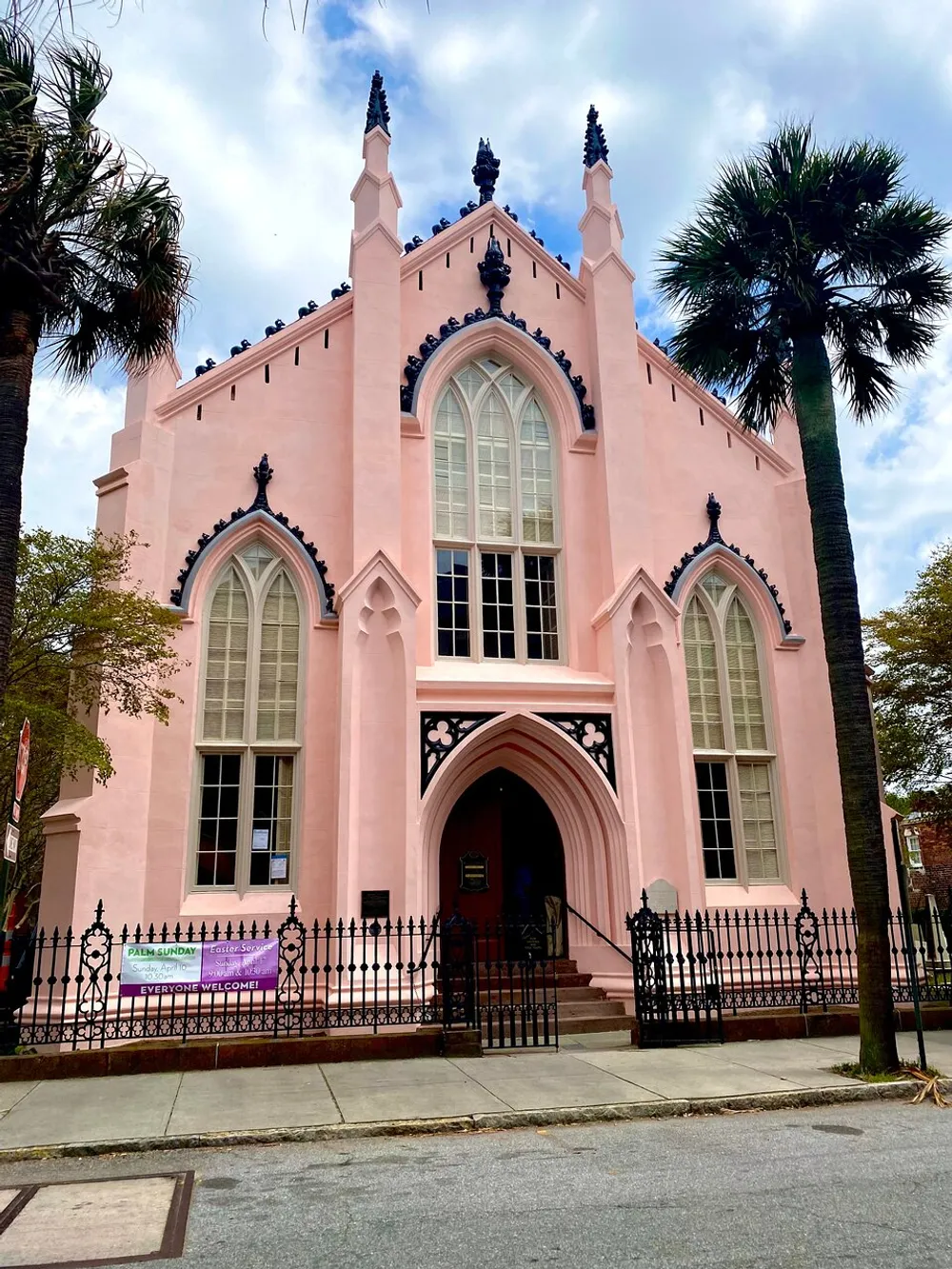 A pastel pink Gothic Revival style church features pointed arch windows and a welcoming banner by the entrance framed by a palm tree and wrought iron fence under a blue sky with scattered clouds