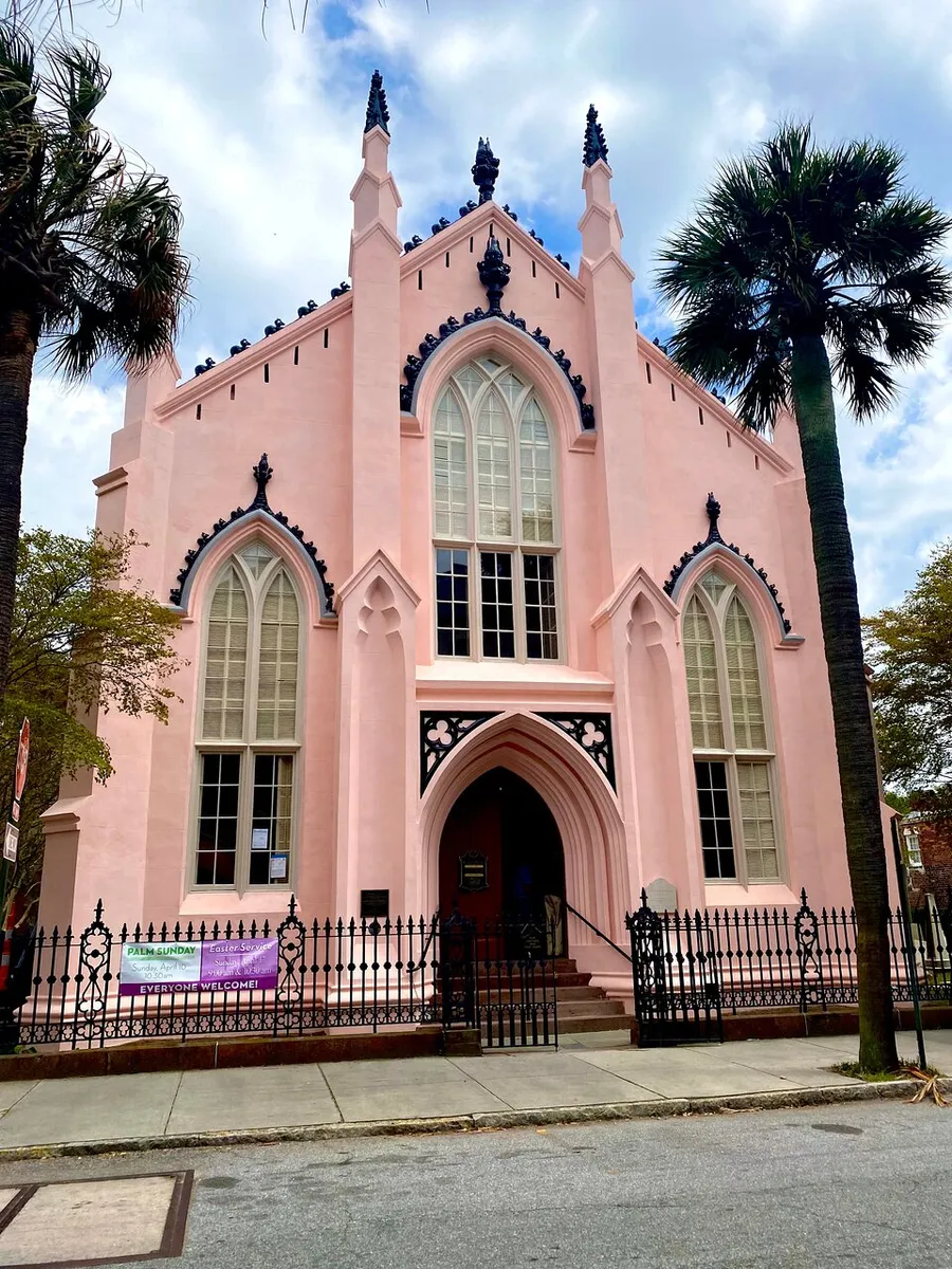 A pastel pink Gothic Revival style church features pointed arch windows and a welcoming banner by the entrance, framed by a palm tree and wrought iron fence under a blue sky with scattered clouds.