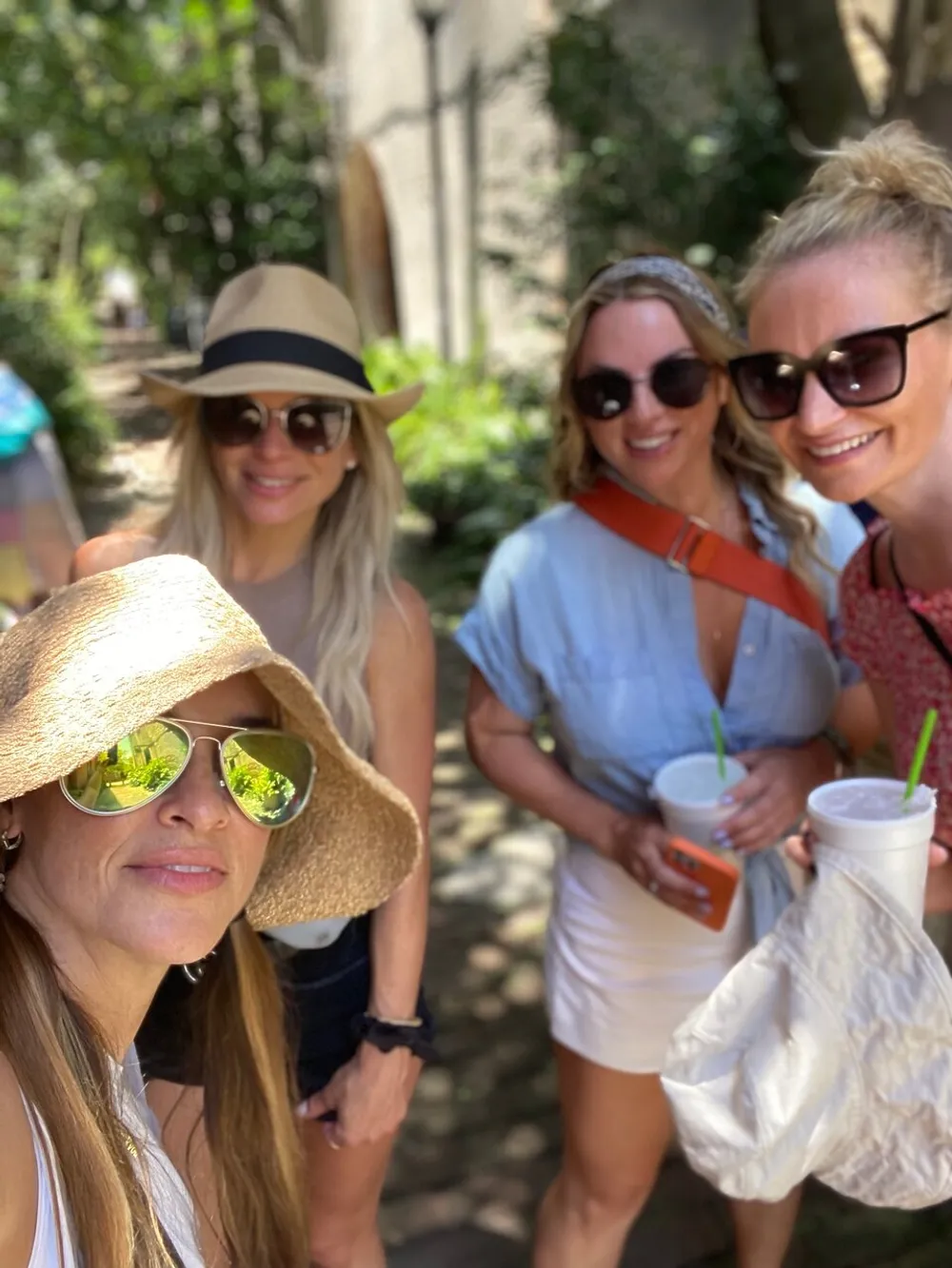 Four smiling women wearing summer attire and sunglasses are posing for a selfie in a sunny outdoor setting with green foliage in the background