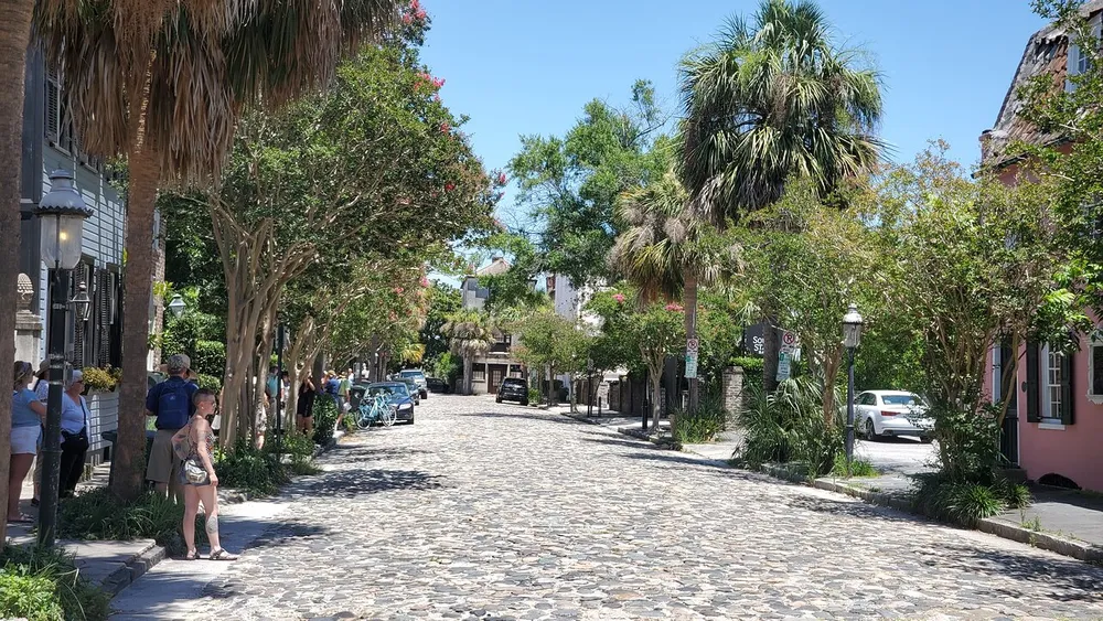 The image captures a sunny day on a charming cobblestone street lined with trees and historical buildings with people walking and enjoying the ambiance
