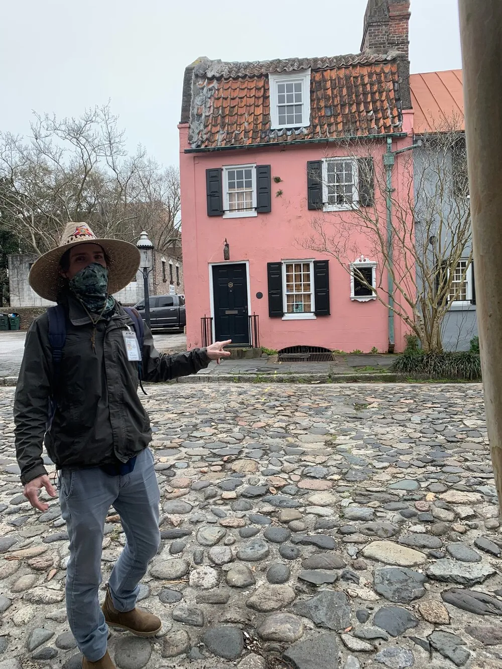A person wearing a hat and mask is gesturing towards a small pink house with a tiled roof on a cobblestone street