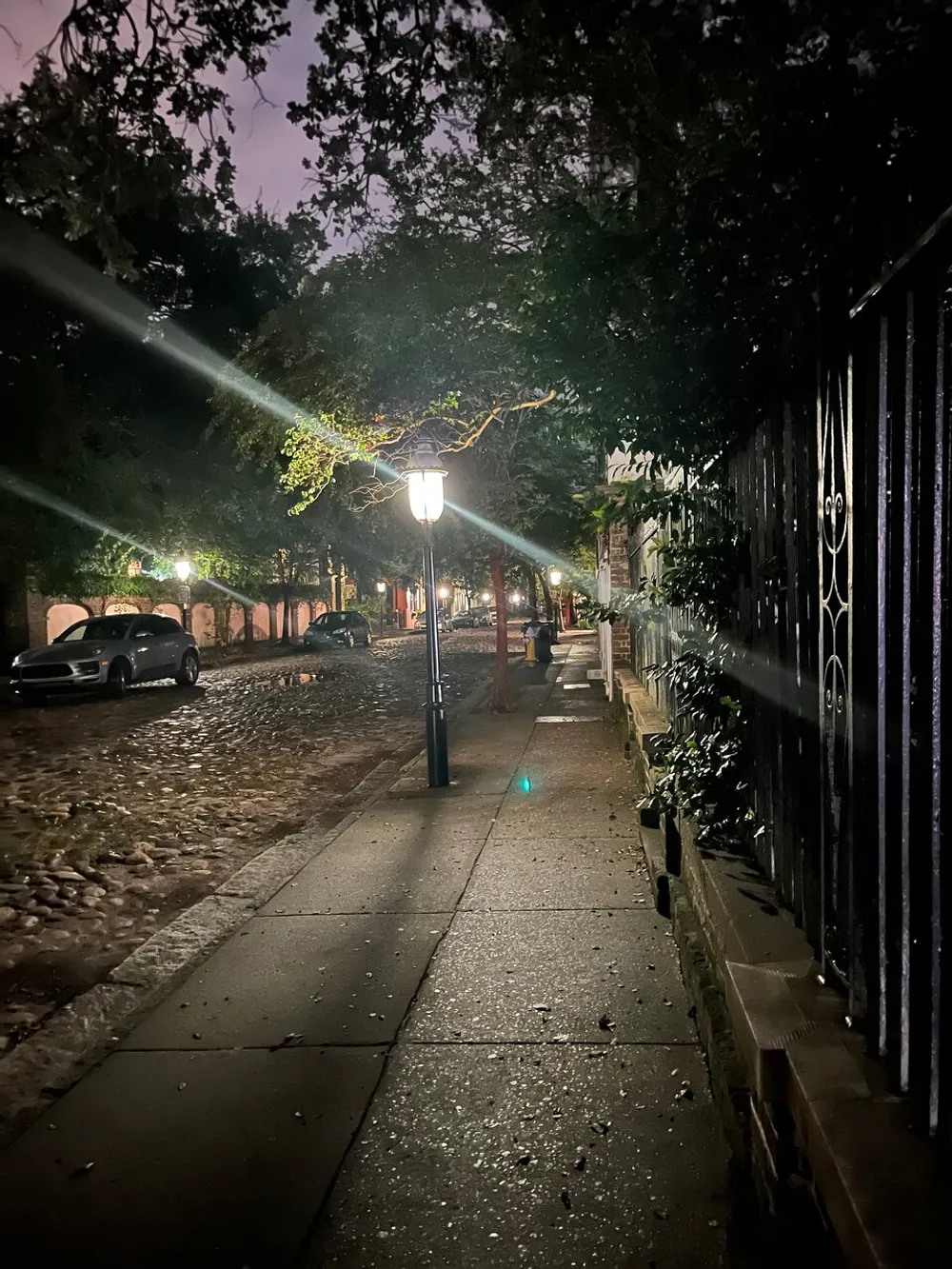 An illuminated streetlight casts a warm glow on a cobblestone-lined sidewalk at dusk creating an atmospheric and serene urban scene
