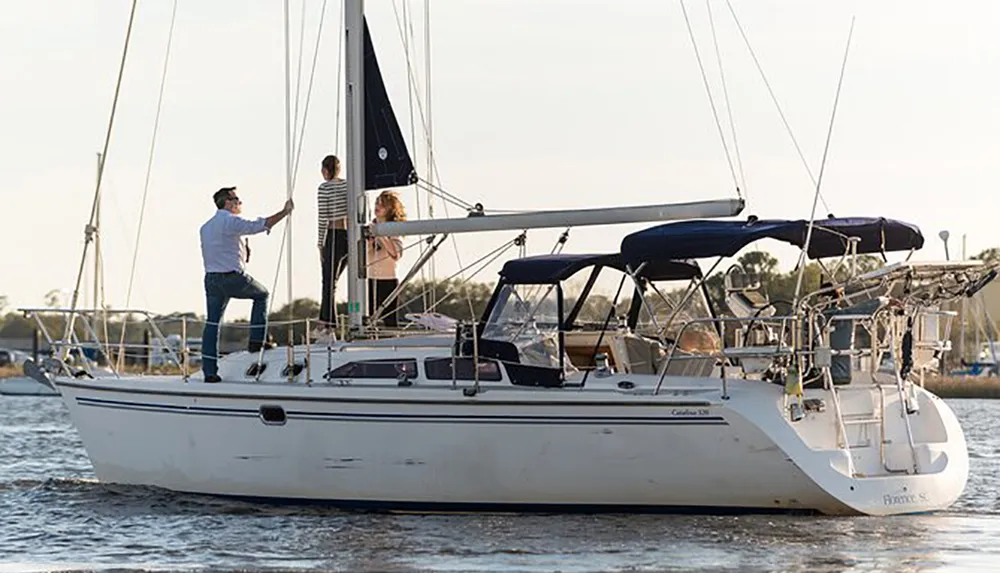 Three people are standing on a sailboat with one of them adjusting the rigging as the boat is docked or sailing near the shore in a calm water setting