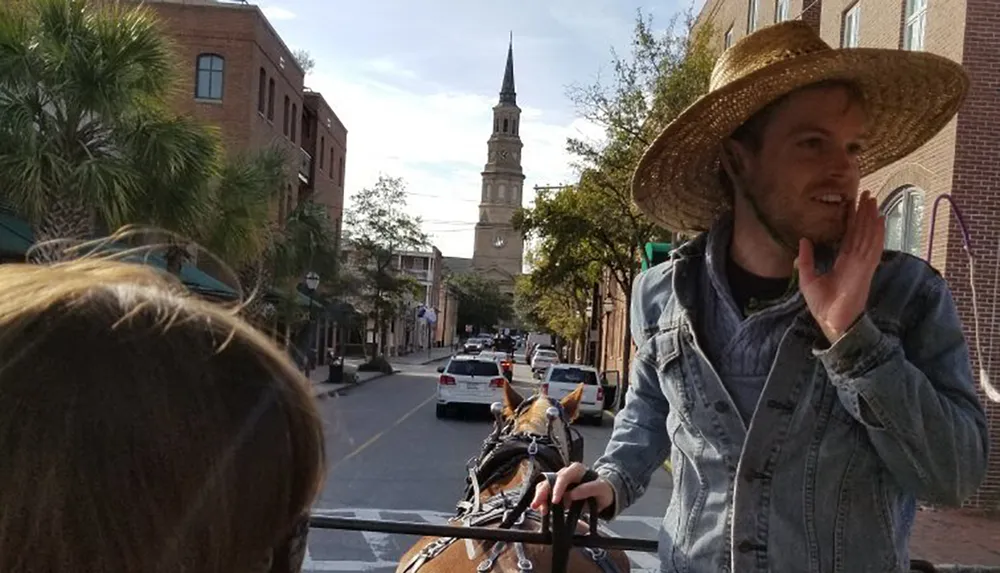 A person in a straw hat is guiding a horse-drawn carriage through a city street with a historic church steeple visible in the background