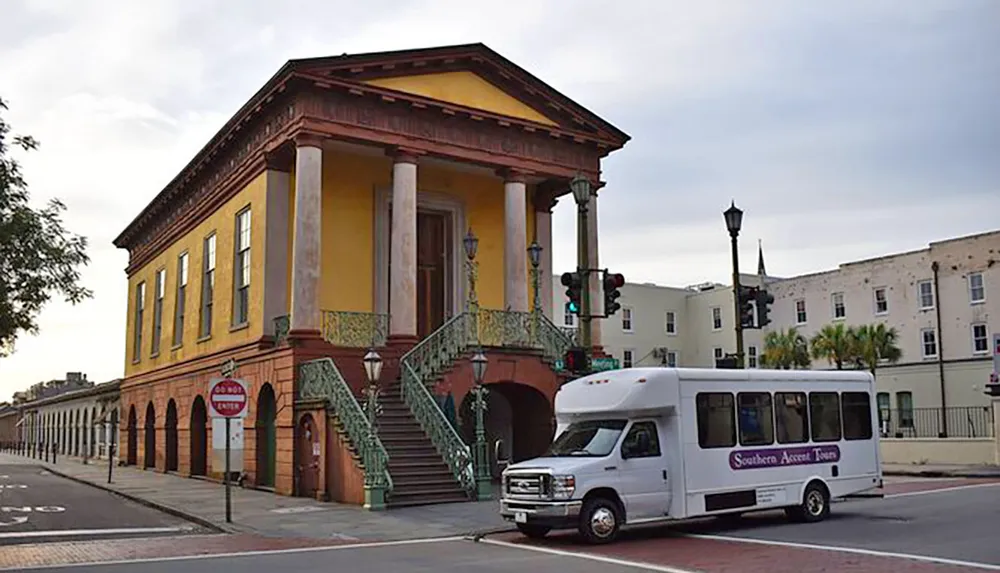 The image shows a historic-looking yellow building with a red roof and green ironwork on the stairs situated on a street corner with a white tour bus passing by