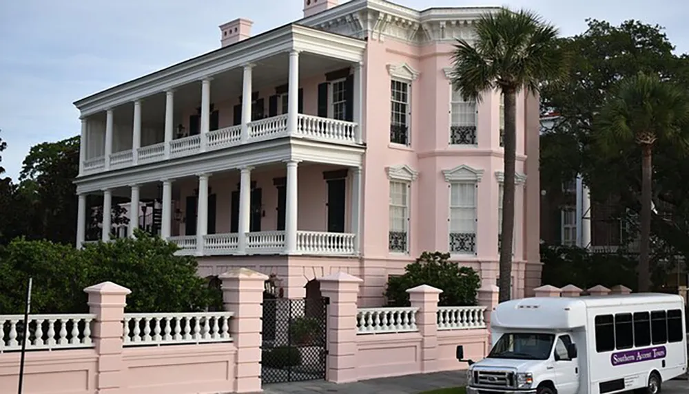 The image shows a striking pink two-story building with white columns and porches surrounded by palm trees and a white fence with a tour bus parked in front
