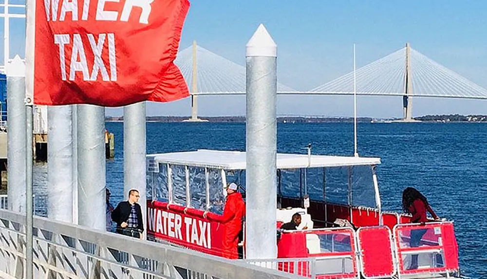 A red water taxi docks at a pier with a WATER TAXI flag on a sunny day with a bridge in the background and a few passengers around