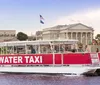 A water taxi with passengers on board is cruising near the shore with a large building in the background