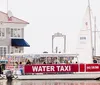 A water taxi with passengers on board is cruising near the shore with a large building in the background