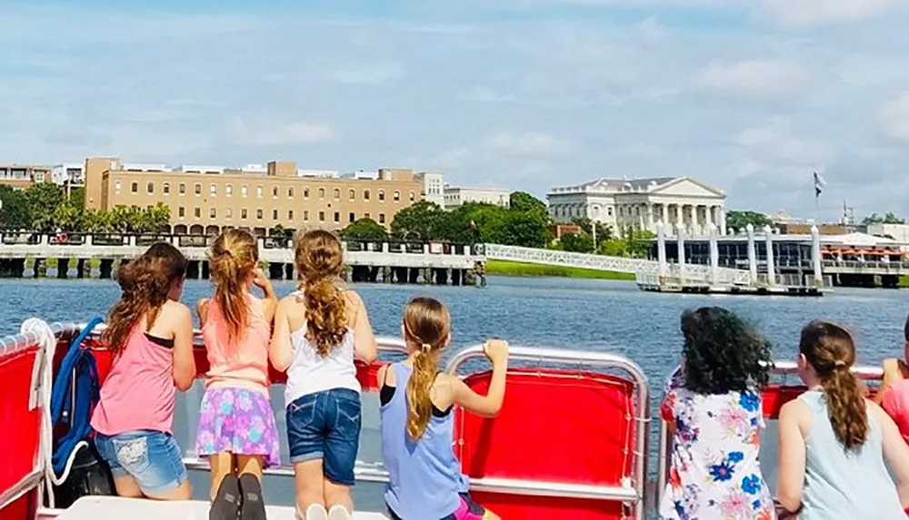 A group of children enjoys a sunny boat tour taking in the views of a waterway with buildings and a bridge in the background