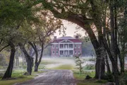 A stately red-brick building stands at the end of a tree-lined gravel path, shrouded in a light morning mist.
