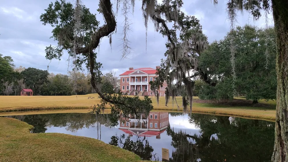 A grand red-brick mansion is framed by Spanish moss-draped trees and reflected in a serene pond under a cloudy sky