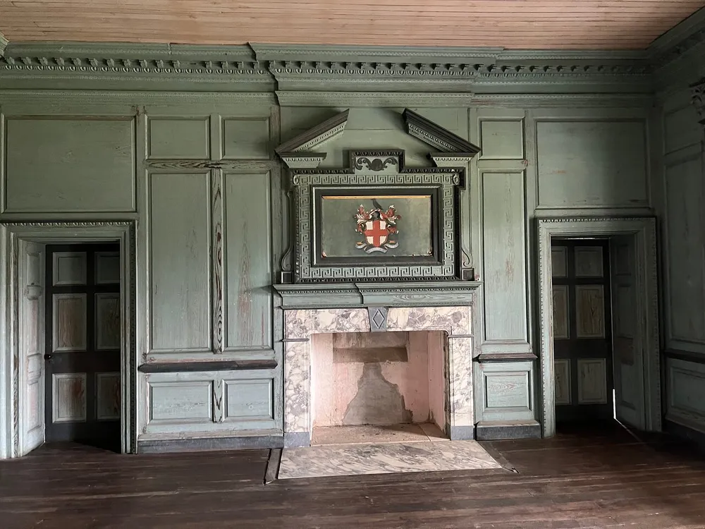 An antique room with ornate green paneling features an elaborate fireplace mantel adorned with a coat of arms flanked by two wooden doors
