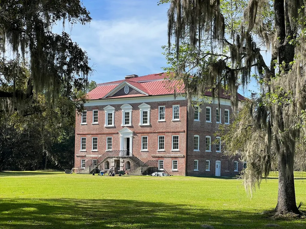 The image features a stately red brick building with white windows and a red tile roof set against a backdrop of lush greenery and hanging Spanish moss from towering trees under a clear blue sky