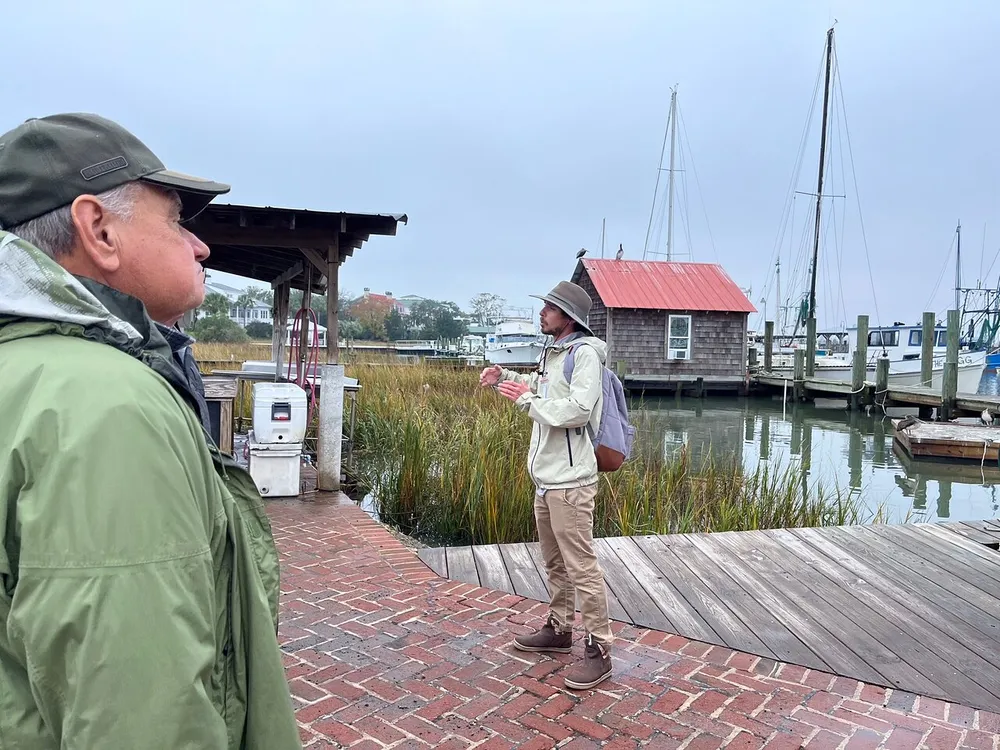 Two men stand on a dock surrounded by sailboats and a waterway seemingly engaged in a conversation in a cloudy tranquil marina setting