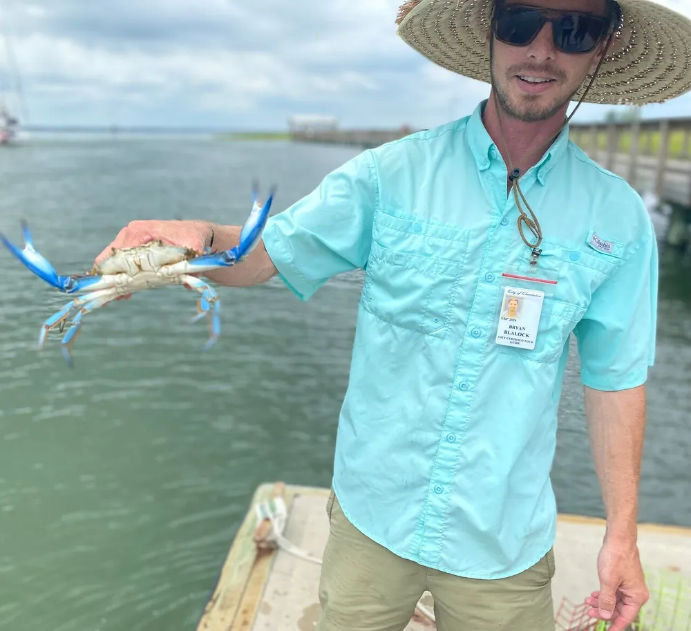 A person wearing a straw hat and a light blue shirt is holding a blue-clawed crab with a dock and water in the background