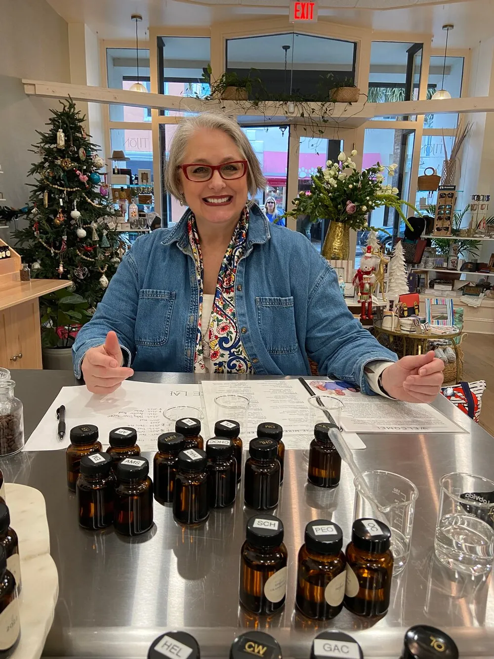 A smiling person is sitting behind a table with an array of small labeled bottles likely in a shop or boutique with a festive atmosphere