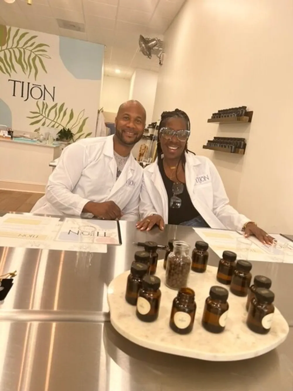 Two smiling individuals in lab coats are seated behind a counter with small bottles of what appear to be fragrance ingredients in a boutique with the name TIJON in the background