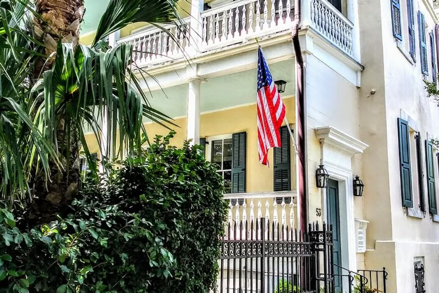 A traditional American flag hangs from the balcony of a charming, pastel-colored house with shutters, nestled amidst lush greenery.