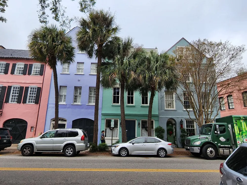 A row of colorful multi-story houses line a street adorned with palm trees and parked vehicles on a cloudy day