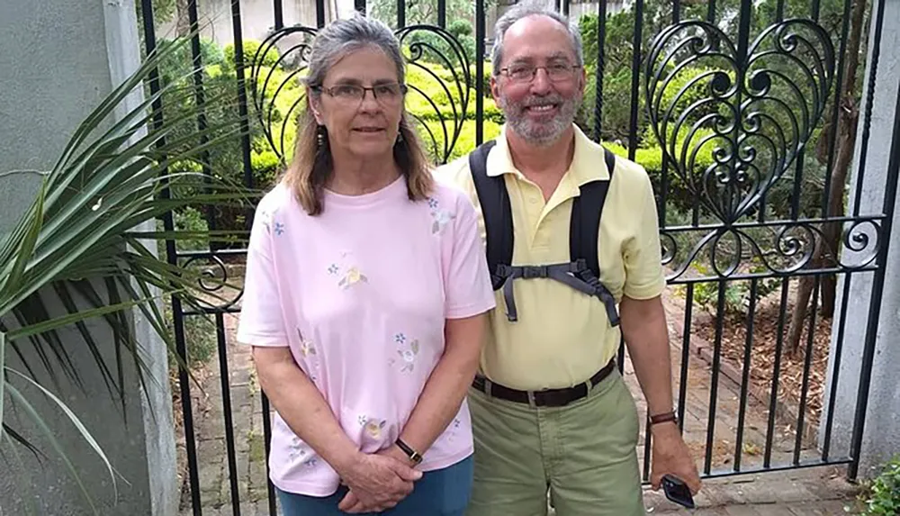 A smiling man with glasses and a beard wearing a yellow shirt and khaki pants with suspenders stands next to a woman in a pink shirt and blue jeans in front of a decorative metal gate surrounded by greenery