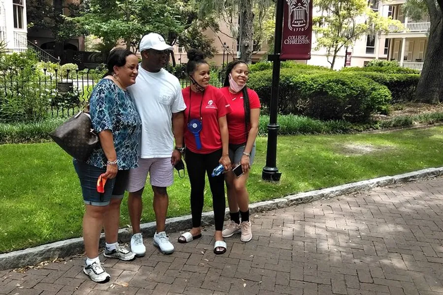 Four people are smiling for a photo on a brick pathway with trees and a lamp post in the background, near a sign that reads College of Charleston.
