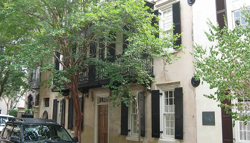 The image shows a quaint multi-story building with a balcony shutters and surrounded by foliage on a sunny day
