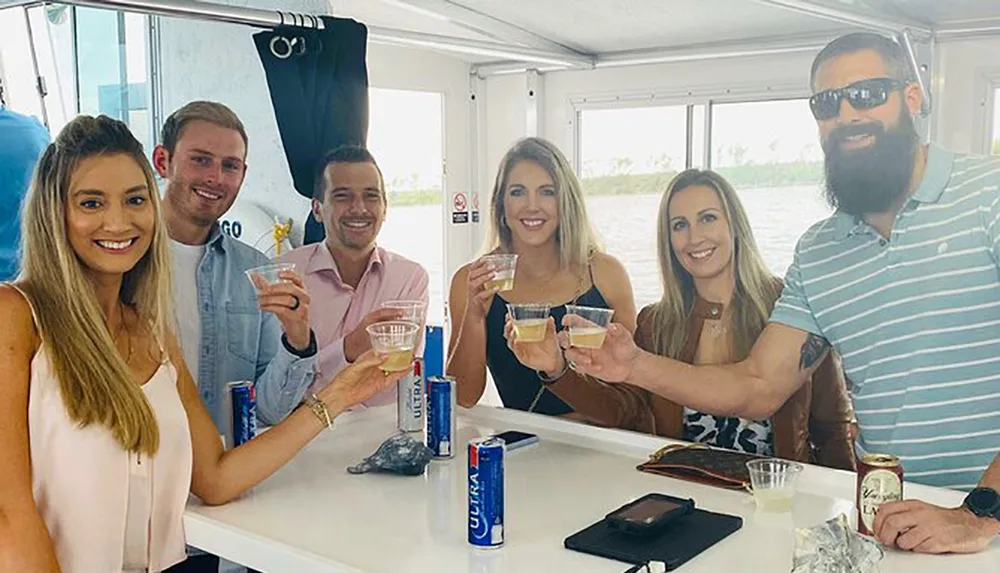A group of six smiling adults are toasting with drinks aboard a boat