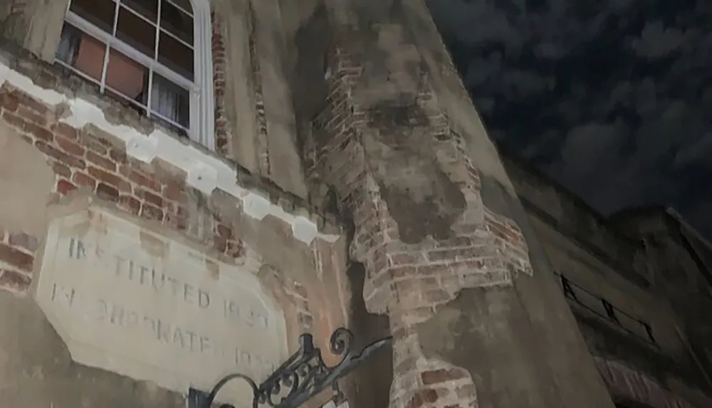 The image shows a night view of a weathered building with exposed bricks and a faded inscription that includes the year 1850 set against a cloudy sky