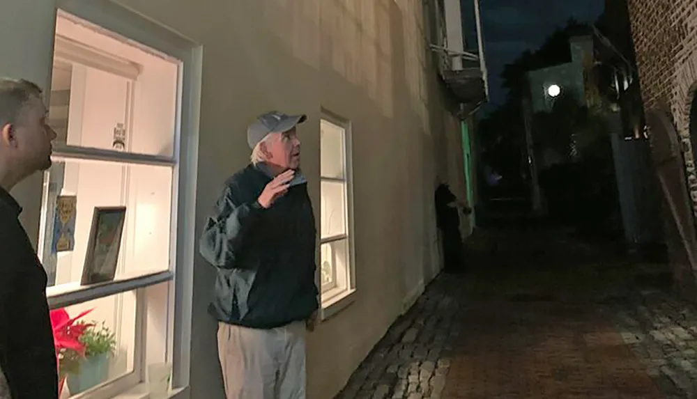 A person is gesturing while speaking to another individual on a cobblestone alleyway at night with a gallery window displaying artwork in the background