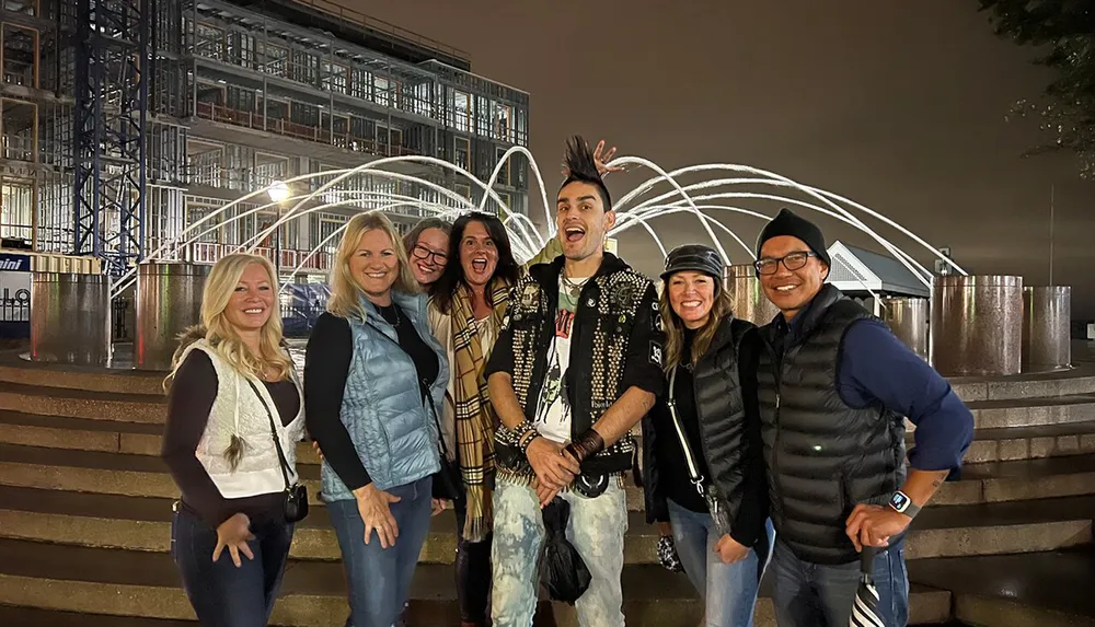 A group of seven people is posing for a photo at night with a fountain in the background and construction scaffolding visible on a building behind them