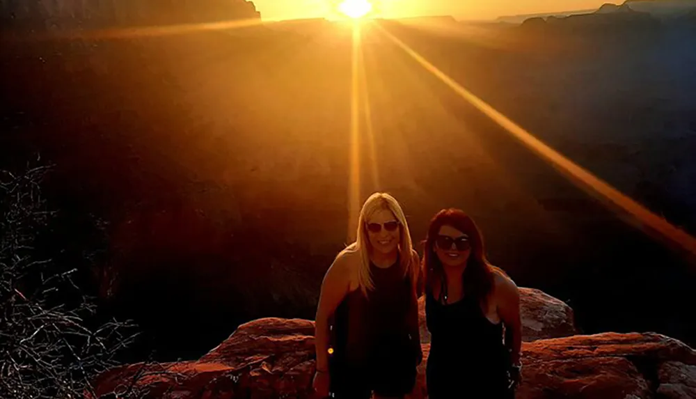 Two people are posing for a photo at sunset with the sun in the background casting rays of light over a scenic canyon