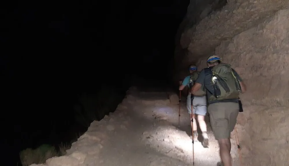 Two hikers are walking along a narrow trail on the side of a steep slope at night using headlamps for guidance