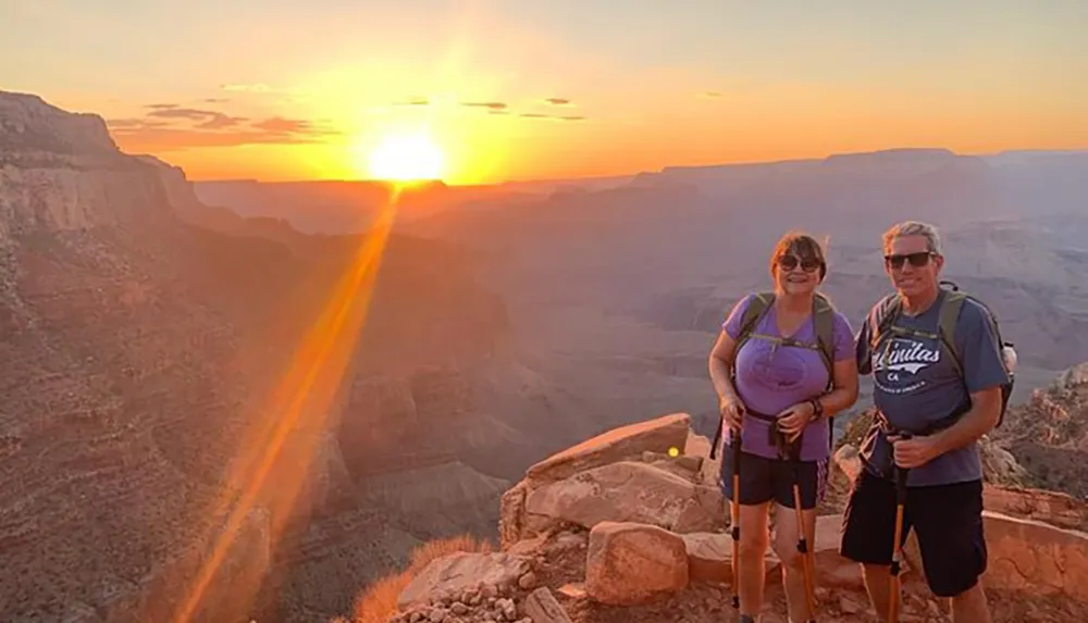 Two hikers are posing for a photo with a breathtaking sunset at the Grand Canyon in the background