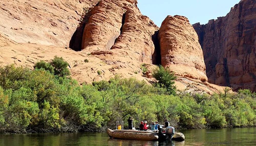 A group of people are on a raft on a calm river with striking red sandstone cliffs in the background