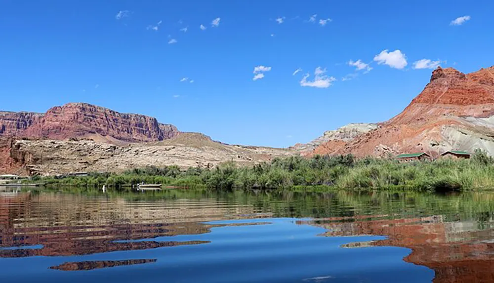 A tranquil river reflects the blue sky and the towering red rock formations on its banks under a partly cloudy sky