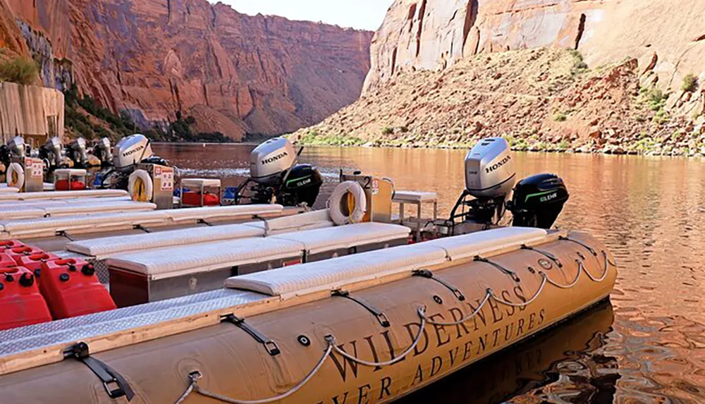 The image shows a motorized rubber raft with Wilderness River Adventures written on the side equipped with several outboard engines and fuel containers docked along a calm river with towering red rock cliffs in the background