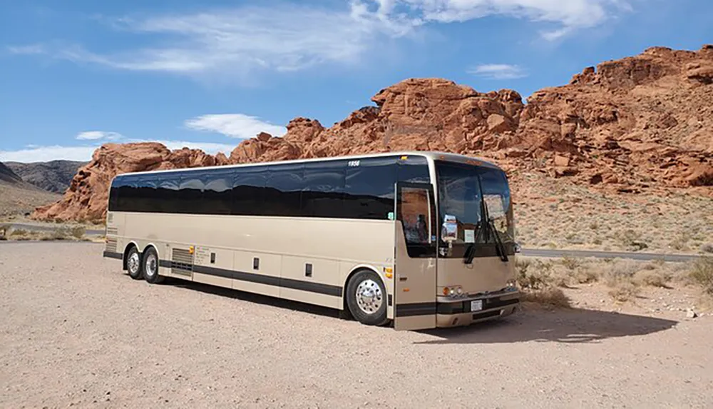 A tour bus is parked on a gravel lot with a backdrop of red rock formations under a bright blue sky