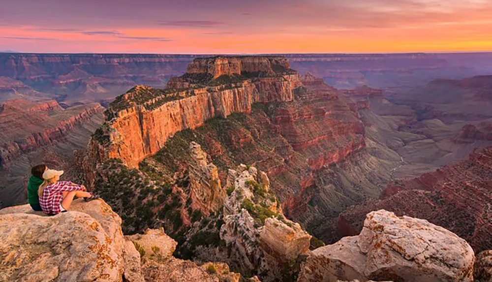 A person sits on a rocky ledge enjoying a colorful sunset over the vast layered rock formations of the Grand Canyon