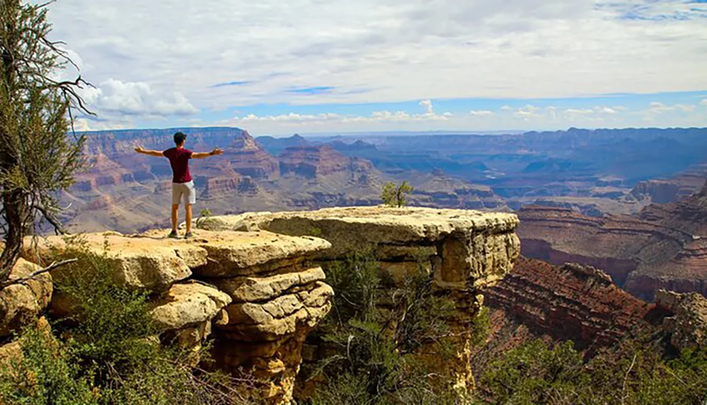 A person stands with outstretched arms on the edge of a cliff overlooking the vast and layered rock formations of the Grand Canyon under a partially cloudy sky