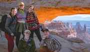 A group of five people pose for a photo under a natural arch with a panoramic view of canyons in the background.