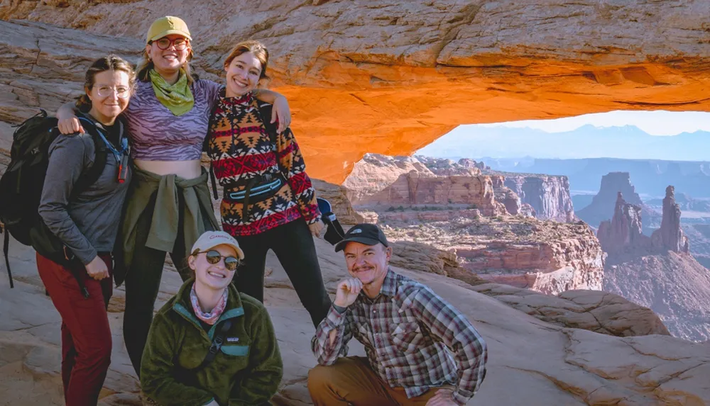 A group of five people pose for a photo under a natural arch with a panoramic view of canyons in the background