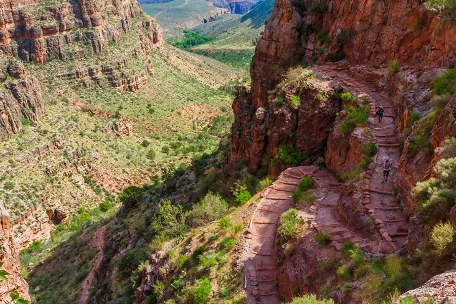 Hikers traverse a steep, winding trail carved into the side of a majestic, red-rock canyon under a bright, clear sky.