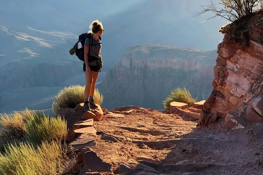 A hiker stands on the edge of a trail overlooking the vast expanse of the Grand Canyon bathed in the warm glow of sunlight.