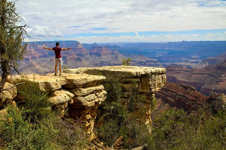 A person with outstretched arms stands on the edge of a rocky cliff overlooking the vast and colorful expanse of the Grand Canyon under a partly cloudy sky.