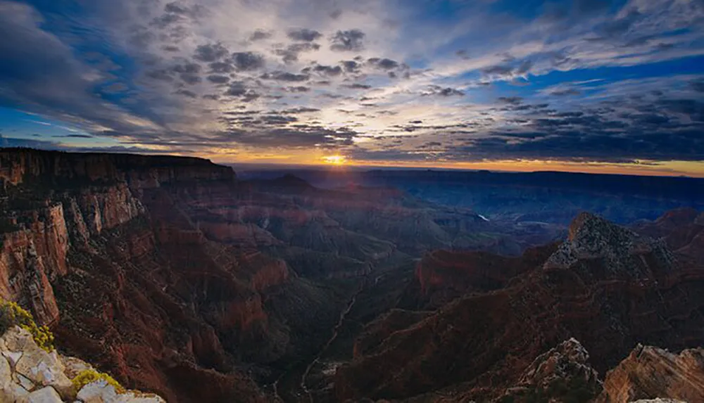 The image captures a breathtaking sunrise over the vast rugged terrain of the Grand Canyon under a dramatic sky