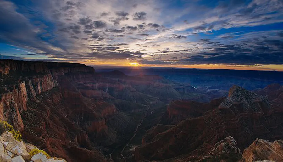 The image captures a breathtaking sunrise over the vast, rugged terrain of the Grand Canyon under a dramatic sky.