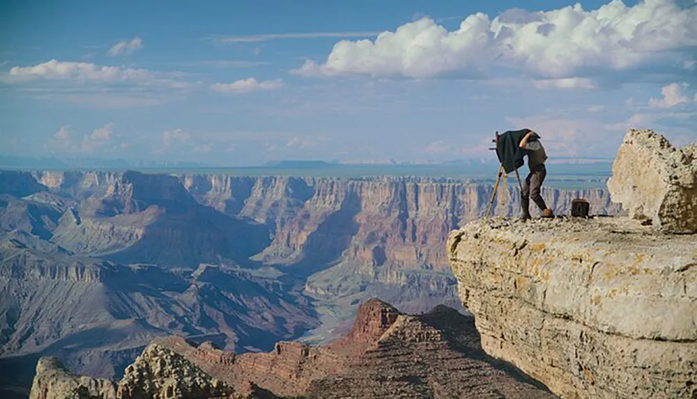 A photographer with a tripod is capturing the vast landscape of the Grand Canyon from a rocky ledge