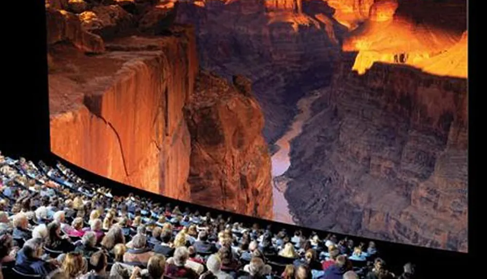 An audience is seated in a theater looking at a large curved screen displaying an image of a grand canyon