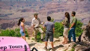 A group of tourists is engaging with a guide near the edge of a canyon, with a pink Jeep Tours vehicle visible in the foreground.